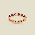 A gold eternity ring featuring a continuous row of square cut dark red gemstones, shown on a solid light beige background.