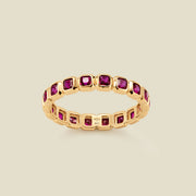A gold eternity ring featuring a continuous row of square cut dark red gemstones, shown on a solid light beige background.