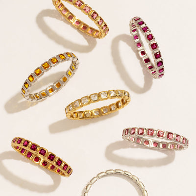 Seven eternity style rings in gold and silver, each with a different color of square cut gemstones in a continuous row, set against a white background with natural shadows.