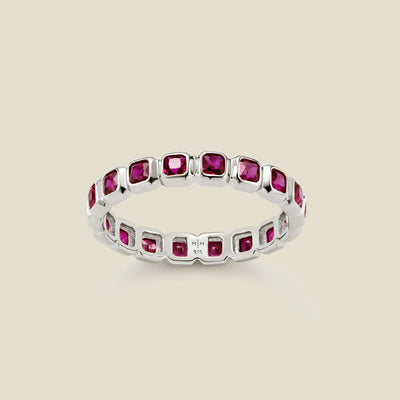 A silver eternity ring featuring a continuous row of square cut dark red gemstones, shown on a solid light beige background.
