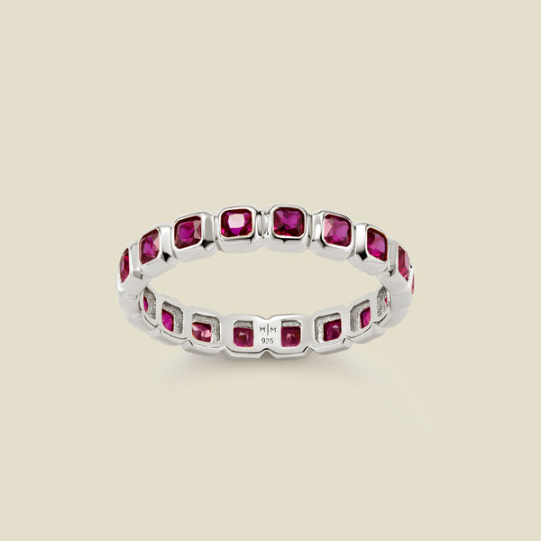 A silver eternity ring featuring a continuous row of square cut dark red gemstones, shown on a solid light beige background.