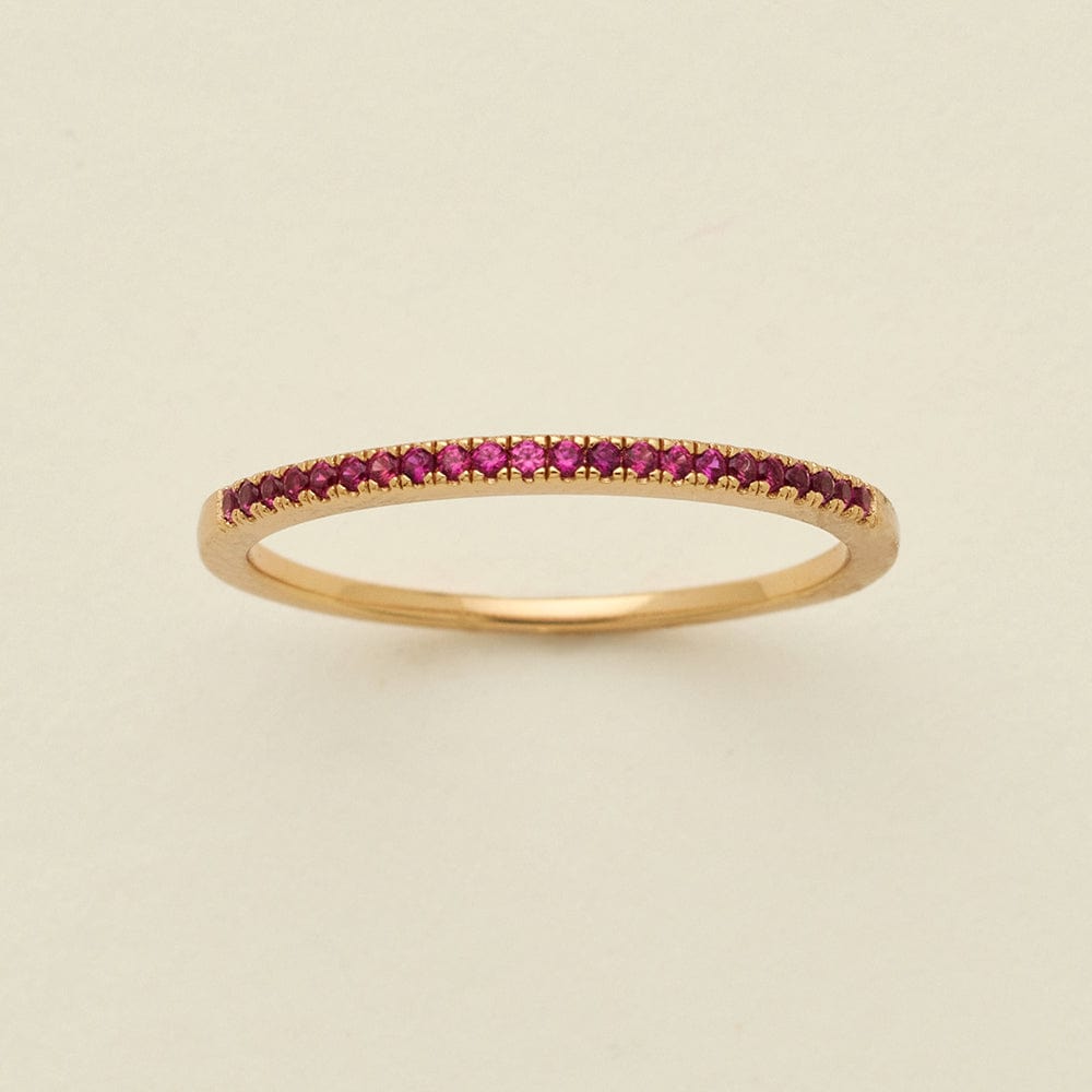A gold ring featuring a single row of small round-cut dark red gemstones, photographed on a clean, solid, warm off-white background.