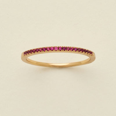 A gold ring featuring a single row of small round-cut dark red gemstones, photographed on a clean, solid, warm off-white background.