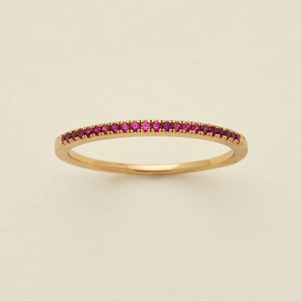 A gold ring featuring a single row of small round-cut dark red gemstones, photographed on a clean, solid, warm off-white background.