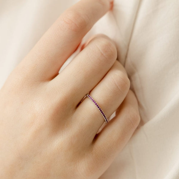 A close-up of a hand resting on a cream cloth with a thin silver ring, featuring a single row of small dark red gemstones across the front.
