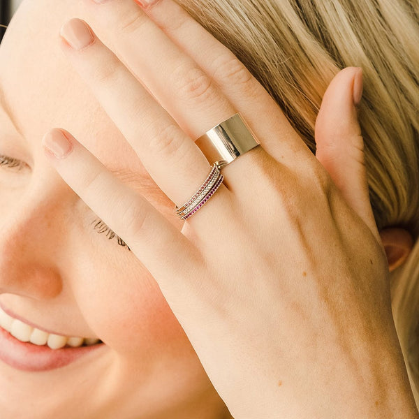 A close-up of a hand resting on a person's face, showcasing a thick gold polished cigar band ring and three thin stacked rings, each with a different color of small gemstones in a single row across the front.