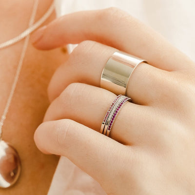 A close-up of a hand showcasing a thick polished gold cigar band ring and a stack of three thin gold rings, each with a different color of small gemstones in a single row across the front.