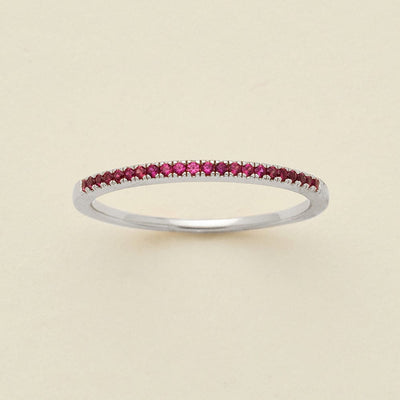 A silver ring featuring a single row of small round-cut dark red gemstones, photographed on a clean, solid, warm off-white background.