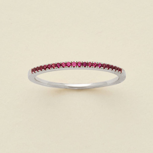A silver ring featuring a single row of small round-cut dark red gemstones, photographed on a clean, solid, warm off-white background.