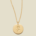 A gold chain necklace with a round gold disc pendant featuring an engraved rose flower design, displayed against a solid light beige background.