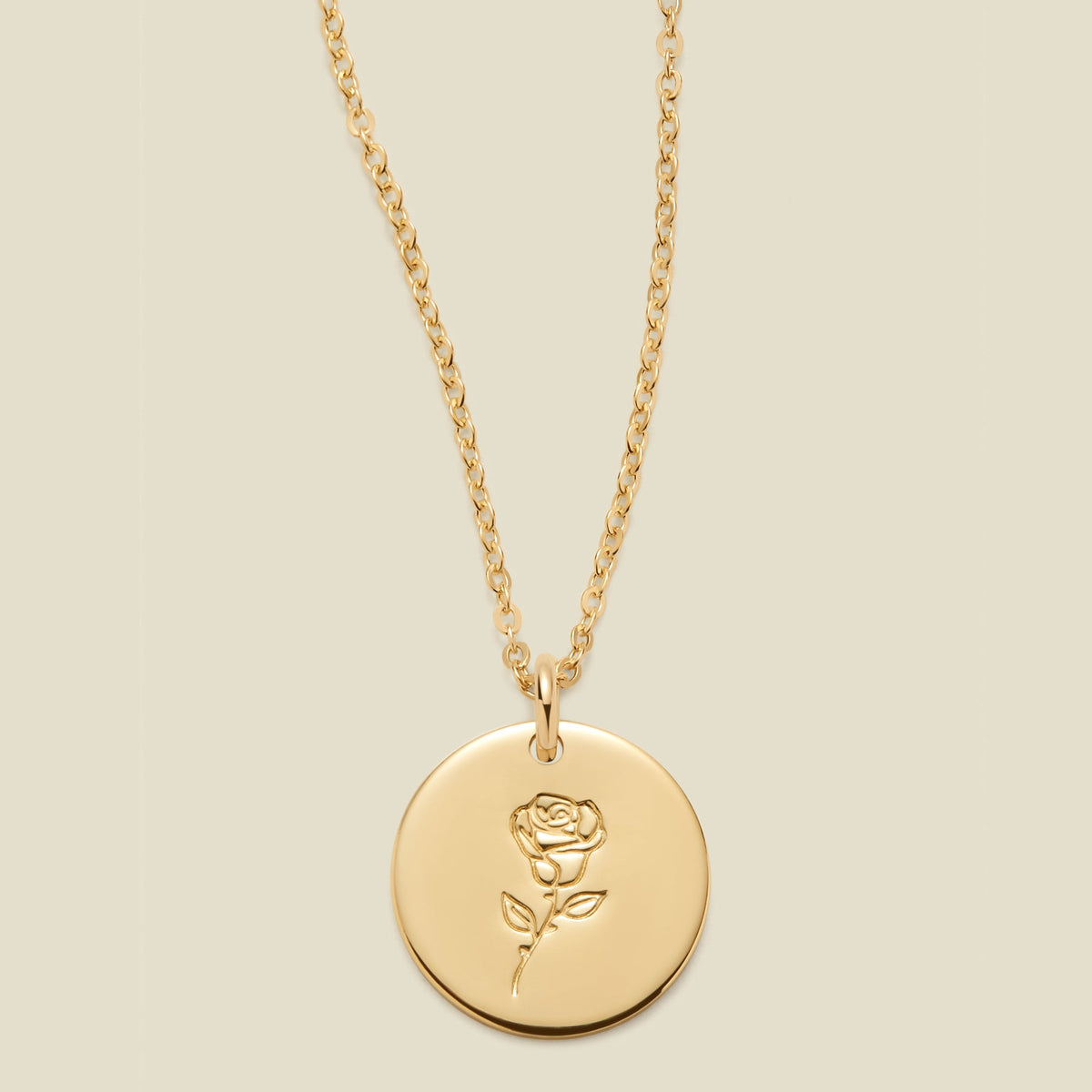 A gold chain necklace with a round gold disc pendant featuring an engraved rose flower design, displayed against a solid light beige background.