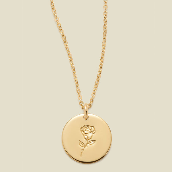 A gold chain necklace with a round gold disc pendant featuring an engraved rose flower design, displayed against a solid light beige background.