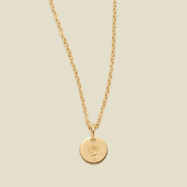 A gold chain necklace with a round gold disc pendant featuring an engraved rose flower design, displayed against a solid light beige background.