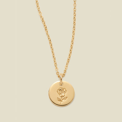 A gold chain necklace with a round gold disc pendant featuring an engraved rose flower design, displayed against a solid light beige background.