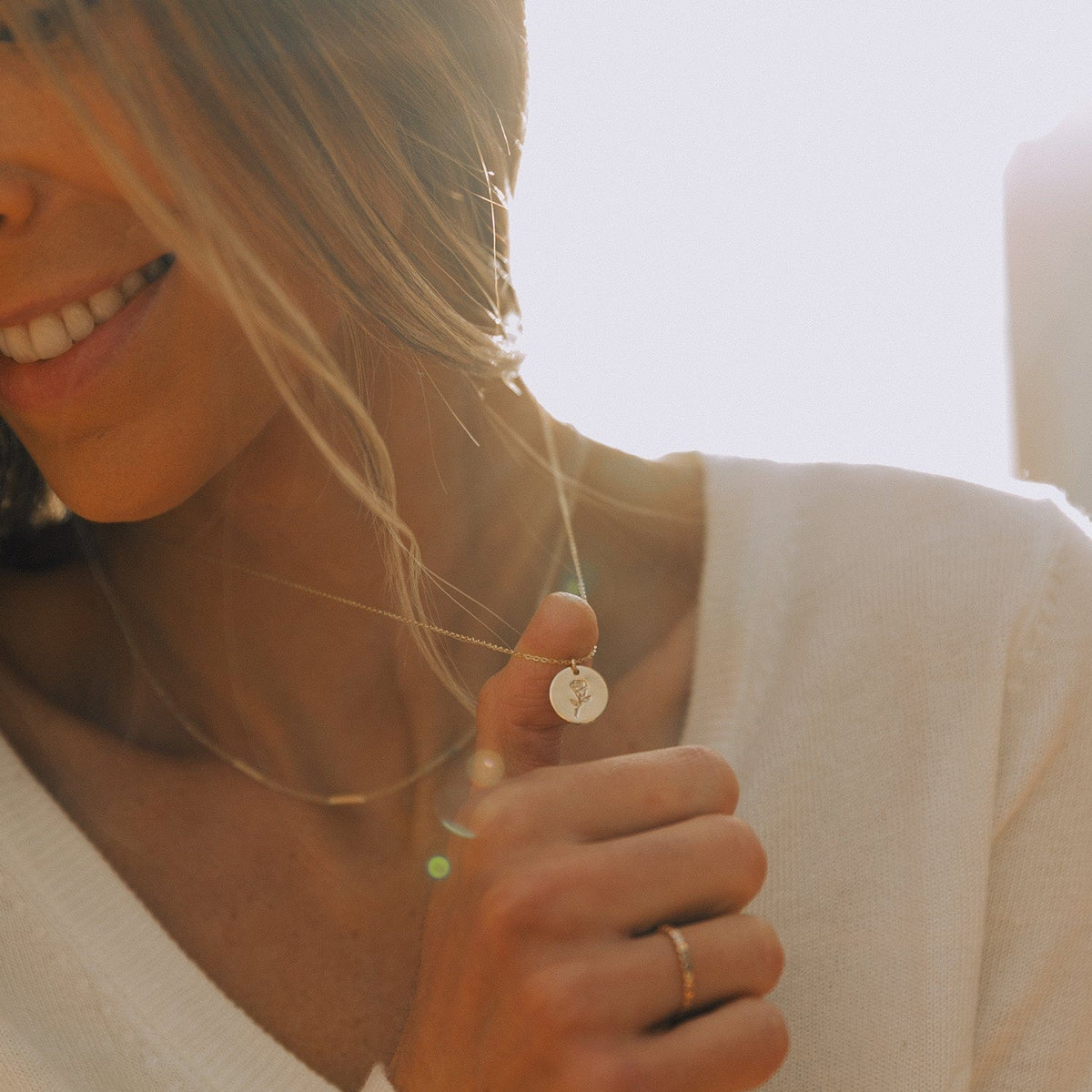 A close-up of a woman in a white knit top with light shining from behind her, holding out a delicate gold pendant chain with a round flat disc charm. The charm is engraved with a rose design.