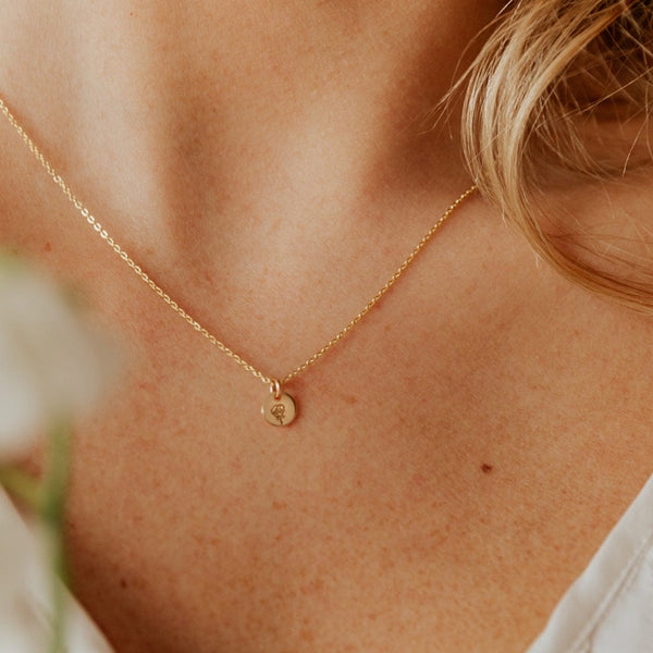 A close-up of a person wearing a delicate gold pendant chain with a small round flat disc charm. The disc is engraved with a rose flower design.