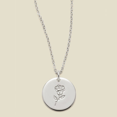 A silver chain necklace with a round silver disc pendant featuring an engraved rose flower design, displayed against a solid light beige background.