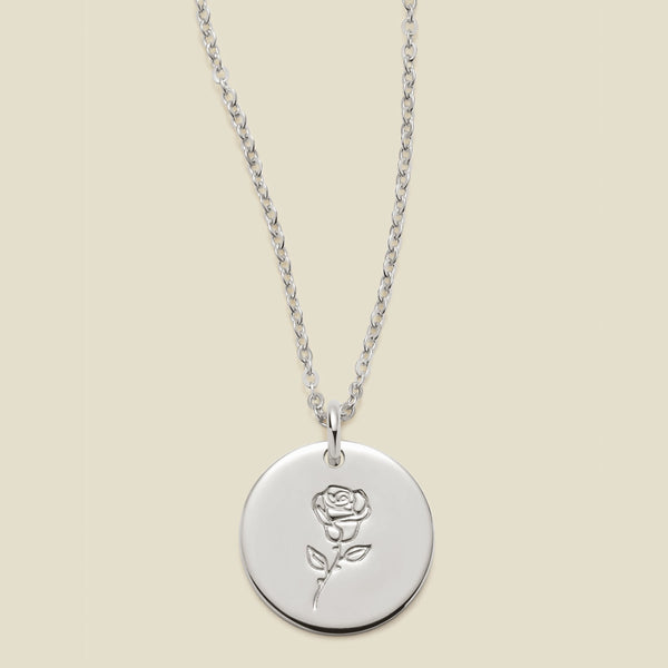 A silver chain necklace with a round silver disc pendant featuring an engraved rose flower design, displayed against a solid light beige background.