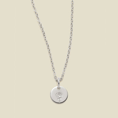 A silver chain necklace with a round silver disc pendant featuring an engraved rose flower design, displayed against a solid light beige background.