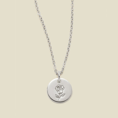 A silver chain necklace with a round silver disc pendant featuring an engraved rose flower design, displayed against a solid light beige background.