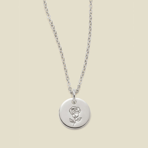 A silver chain necklace with a round silver disc pendant featuring an engraved rose flower design, displayed against a solid light beige background.