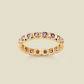 A gold eternity ring featuring a continuous row of square cut light purple gemstones, shown on a solid light beige background.