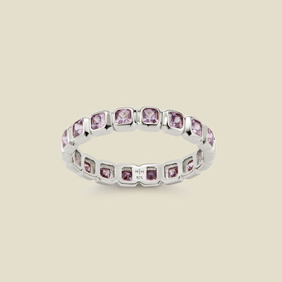 A silver eternity ring featuring a continuous row of square cut light purple gemstones, shown on a solid light beige background.