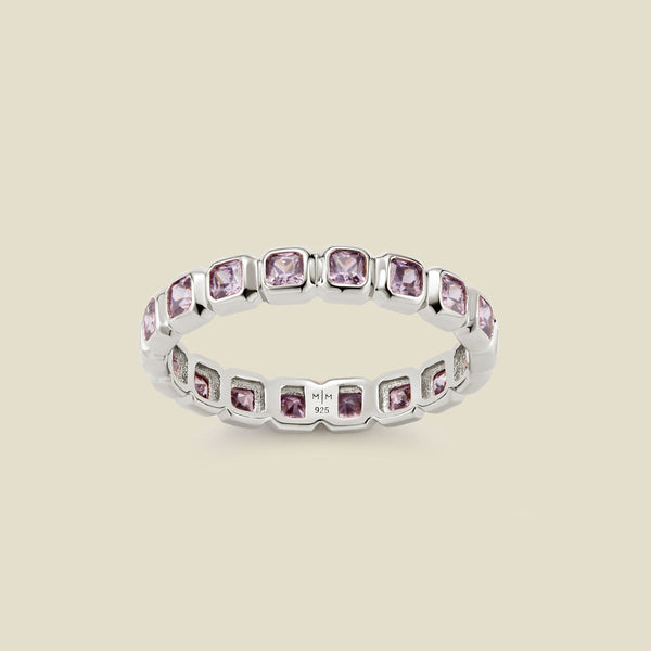 A silver eternity ring featuring a continuous row of square cut light purple gemstones, shown on a solid light beige background.