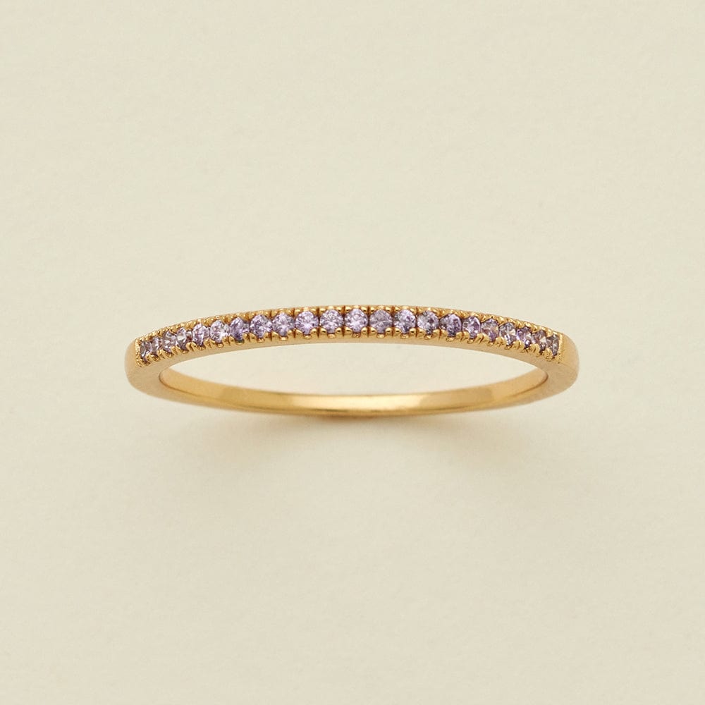 A gold ring featuring a single row of small round-cut light purple gemstones, photographed on a clean, solid, warm off-white background.