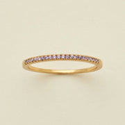 A gold ring featuring a single row of small round-cut light purple gemstones, photographed on a clean, solid, warm off-white background.