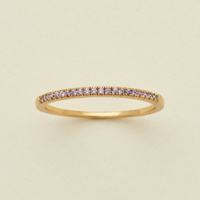 A gold ring featuring a single row of small round-cut light purple gemstones, photographed on a clean, solid, warm off-white background.