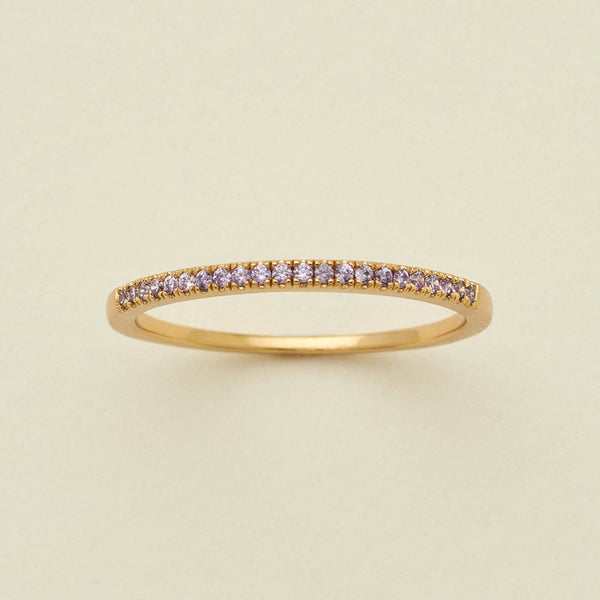 A gold ring featuring a single row of small round-cut light purple gemstones, photographed on a clean, solid, warm off-white background.