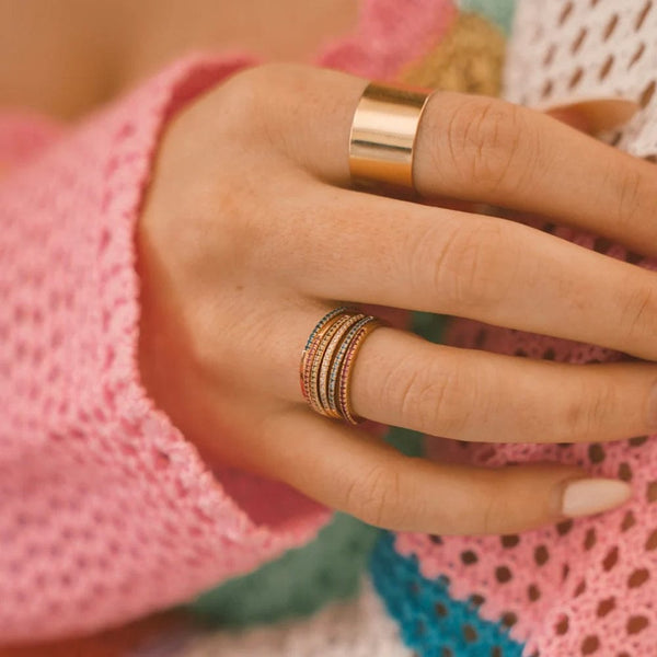 A close-up of a hand resting on a pink, blue, and white open knit sweater, showcasing a thick polished gold cigar band ring and a stack of multiple thin gold rings, each with a different color of small gemstones in a single row across the front.