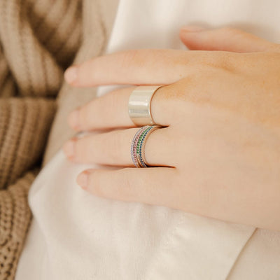 A close-up of a hand resting on a white linen top, showcasing a thick gold polished cigar band ring and a stack of four thin gold rings, each with a different color of small gemstones in a single row across the front. The gemstone colors are light purple, red, green, and blue.