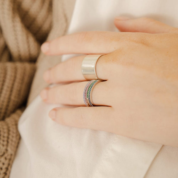 A close-up of a hand resting on a white linen top, showcasing a thick gold polished cigar band ring and a stack of four thin gold rings, each with a different color of small gemstones in a single row across the front. The gemstone colors are light purple, red, green, and blue.
