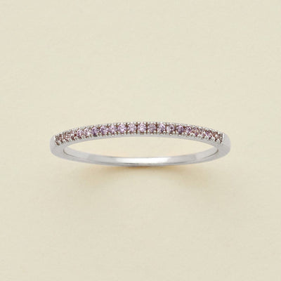 A silver ring featuring a single row of small round-cut light purple gemstones, photographed on a clean, solid, warm off-white background.