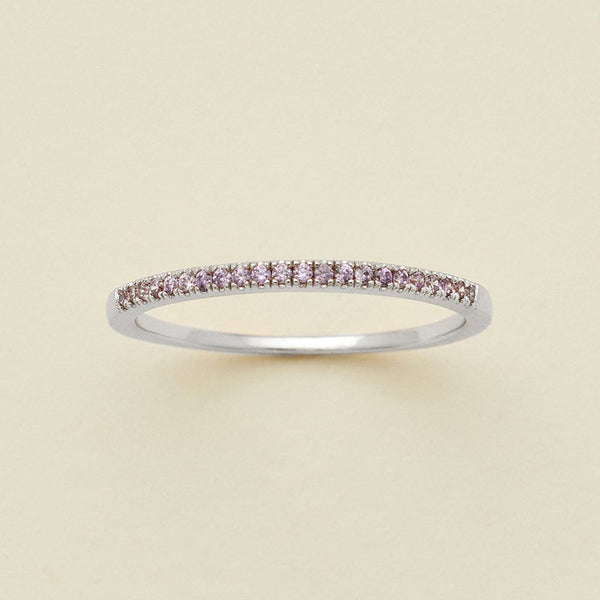 A silver ring featuring a single row of small round-cut light purple gemstones, photographed on a clean, solid, warm off-white background.