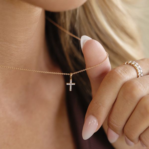 A close-up lifestyle image of a person wearing a gold chain necklace with a small cross charm with clear gemstones, and two gold rings.
