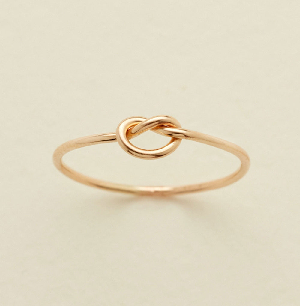 A delicate gold ring featuring a central knotted loop design, photographed on a clean, solid, light beige background.