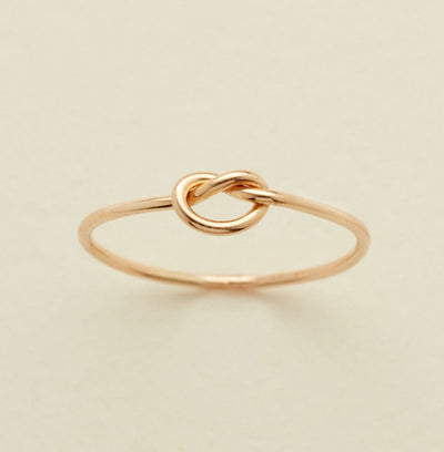 A delicate gold ring featuring a central knotted loop design, photographed on a clean, solid, light beige background.