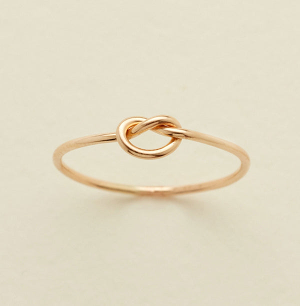 A delicate gold ring featuring a central knotted loop design, photographed on a clean, solid, light beige background.