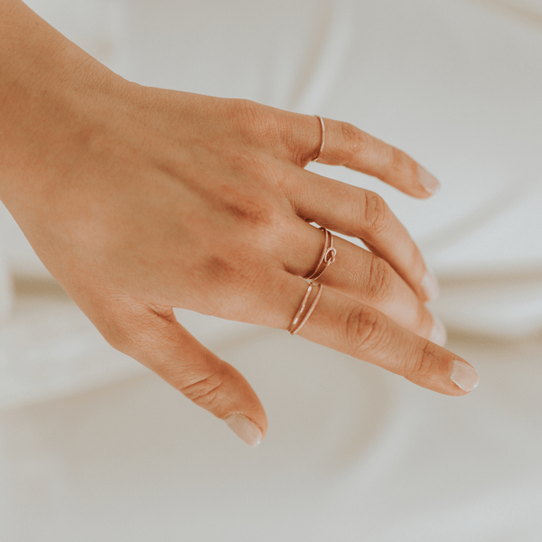 A person's hand in front of a white surface, showcasing multiple thin gold wire rings including one with a knot shape in the center.