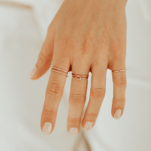 A person's hand in front of a white surface, showcasing multiple thin gold wire rings including one with a knot shape in the center.