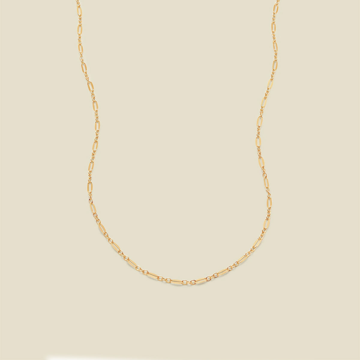 A delicate gold chain necklace with elongated oval links, displayed against a solid, warm beige background.