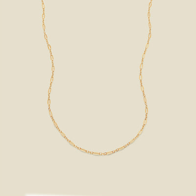 A delicate gold chain necklace with elongated oval links, displayed against a solid, warm beige background.