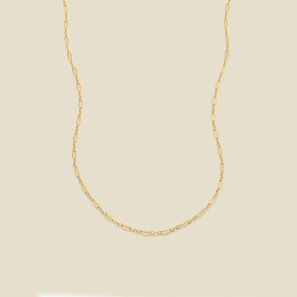 A delicate gold chain necklace with elongated oval links, displayed against a solid, warm beige background.
