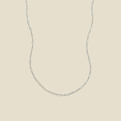A delicate silver chain necklace with elongated oval links, displayed against a solid, warm beige background.