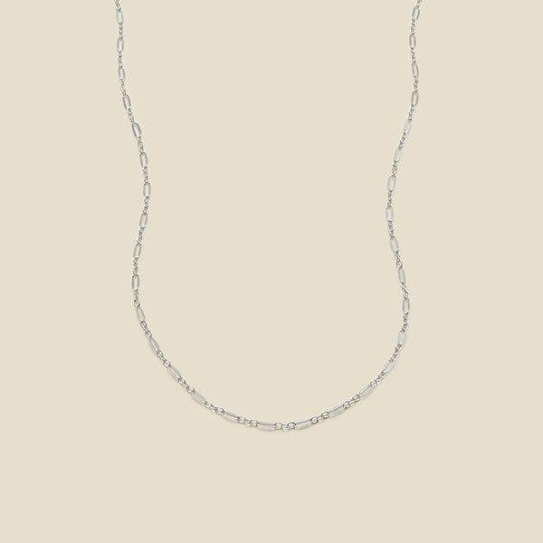 A delicate silver chain necklace with elongated oval links, displayed against a solid, warm beige background.