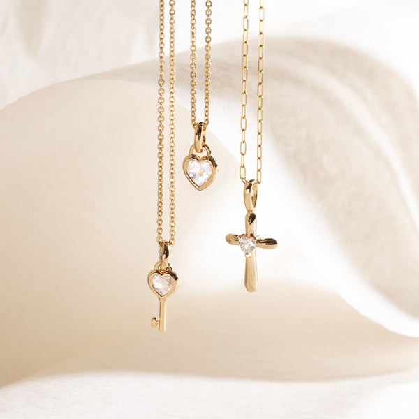 Three gold pendant necklaces with heart, key, and cross pendants on a white background