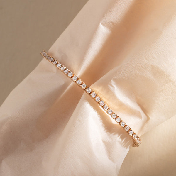 A gold prong-set tennis bracelet wrapped around a cream colored tissue paper, set against a warm beige background.